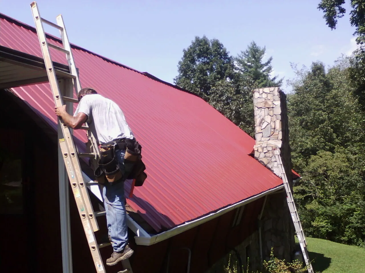 Roofer on a ladder beside a red metal roof and stone chimney — Soffit & Fascia Repair by Roofing Brothers in Brandon